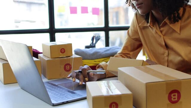 African young woman owner of a start up business designing and selling clothes online checking orders on laptop and preparing for delivery.