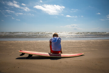 Young male child at the beach sitting on a surfboard with waves and sky in the background
