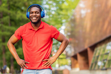 Fototapeta na wymiar african american man in red t-shirt listening music in blue headphones and being in high spirit outdoors