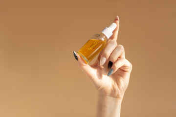 Glass bottle with orange cosmetic serum in woman's hand on brown background. Mockup of container...