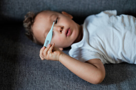 Sick Toddler Having Fever Holding Digital Electronic Thermometer Measuring Temperature While Lying On Sofa At Home, Playing With Medical Equipment. Cold And Flu Symptoms In Children