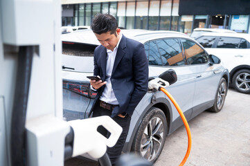 Asian businessman traveling with electric car stopping at charging station standing plugged in internet cable on smartphone smiling joyfully while charging, energy saving electric car view