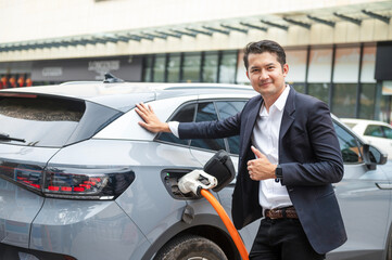 Asian businessman traveling with electric car stopping at charging station standing plugged in internet cable on smartphone smiling joyfully while charging, energy saving electric car view