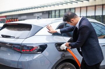 Asian businessman traveling with electric car stopping at charging station standing plugged in internet cable on smartphone smiling joyfully while charging, energy saving electric car view