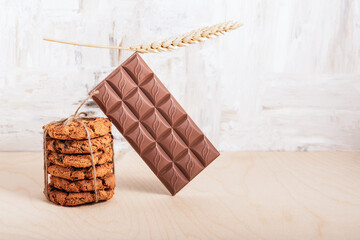 A stack of chocolate chip cookies tied with twine, a bar of chocolate and a spikelet on a wooden table.