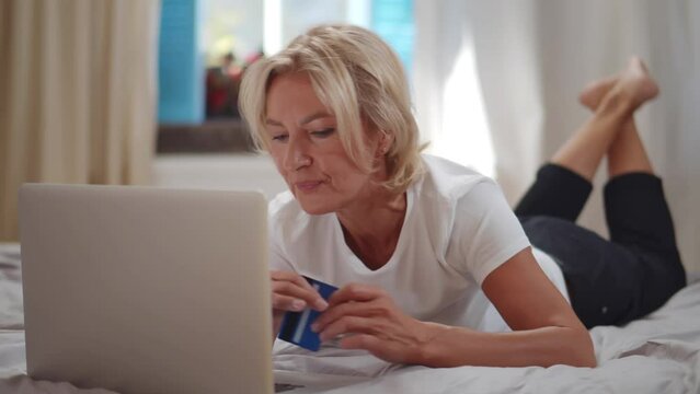 Mature woman making online payment holding bank card using modern laptop at home. Realtime