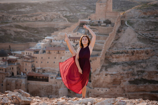 A beautiful brunette girl in a red silk dress dances opposit Mar Saba Monastery located in Jericho in the middle of Negev desert and looks at sunset. Exotic holiday vacation