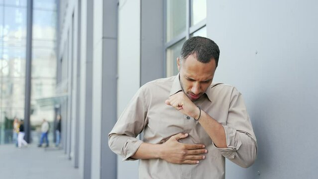 Young adult man is coughing while standing outside an office building. Sick male of mixed race suffers from pneumonia or inflammation of the lungs He is unwell, has a fever, a cold, a virus or the flu