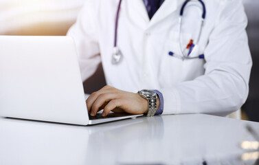 Fototapeta premium Unknown male doctor sitting and working with laptop in a darkened clinic, glare of light on the background, close-upof hands