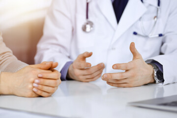 Unknown male doctor and patient woman discussing something while sittingin a darkened clinic, glare of light on the background. Close-up of hands