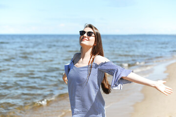 Happy smiling woman in free bliss on ocean beach standing with open hands. Portrait of a brunette female model in summer dress enjoying nature during travel holidays vacation outdoors