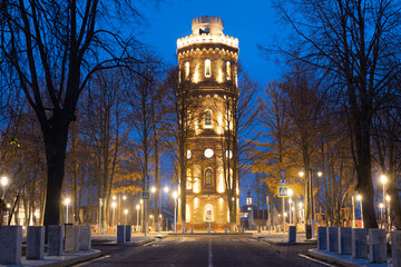 Fototapeta na wymiar Historic brick water tower built in 1916 in the town of Zaraysk, Moscow region, Russia. View on old water tower against the backdrop of an illuminated evening street with golden lanterns.