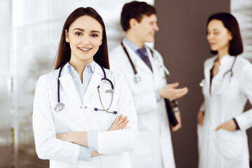 Fototapeta premium Smiling intelligent woman-doctor is standing with arms crossed in a clinic, together with her colleagues at the background. Portrait of physicians at work