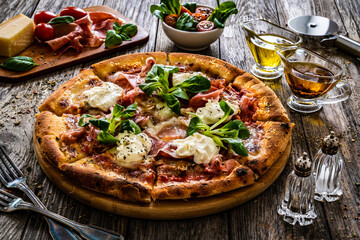 Circle prosciutto pizza with mascarpone cheese and leafy greens on wooden table
