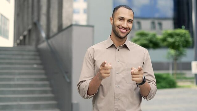 Handsome young adult man pointing fingers of both hands at the camera and showing a like gesture while standing on the street near an office building. A mixed race businessman in a shirt is smiling