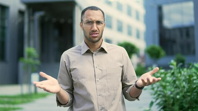 Surprised puzzled young male spreads his hands, shrugs his shoulders, does not understand what happened, looking at the camera while standing on the street. Man has a confused expression on his face
