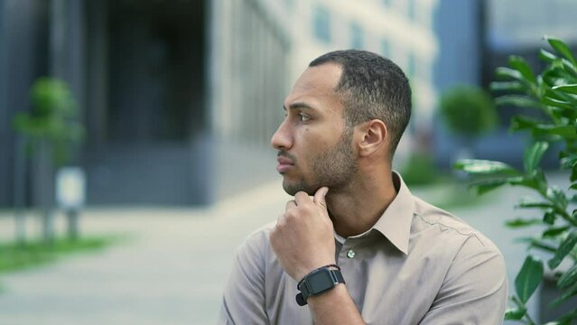 Side view. Close up portrait of a young thoughtful man in a shirt sitting on a bench on the street near an office building. Serious handsome mixed race male looking away while waiting to meet