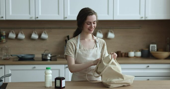 Young woman in apron unpack bag with diverse grocery and dairy products ready for dinner preparation standing in domestic kitchen. Client of delivery services, supermarket webstore customer, cuisine