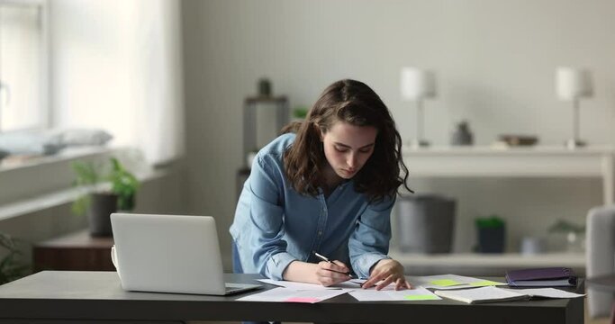 Focused young 25s woman working or studying, prepare task, makes assignment, holds pen writes down information, thoughts, solutions, do essay or summary leaned at desk. Workflow, education, paperwork
