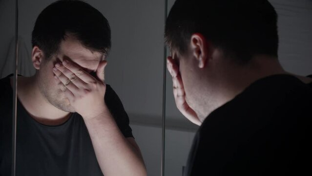 Upset and stressed man standing at mirror and covering face with hands. Concept of depression, suicide, stress, mental illness, loneliness and frustration