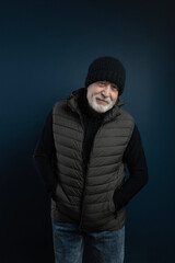 Fototapeta na wymiar Charismatic elderly gray-haired man in hat and warm jacket posing for camera in studio, standing and looking with smile, medium close-up, blue background