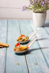 Appetizers of potato salad, tomato, green beans, egg and tuna in ceramic spoons on a wooden background.