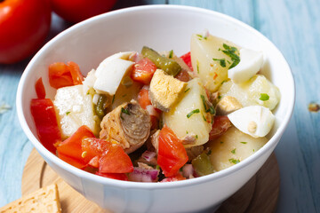 Close-up of  potato salad, tomato, egg, green beans, onion, parsley and tuna in a white bowl.