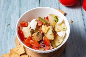 Close-up of  potato salad, tomato, egg, green beans, onion, parsley and tuna in a white bowl.