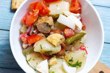 Close-up of  potato salad, tomato, egg, green beans, onion, parsley and tuna in a white bowl.