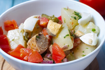 Close-up of  potato salad, tomato, egg, green beans, onion, parsley and tuna in a white bowl.