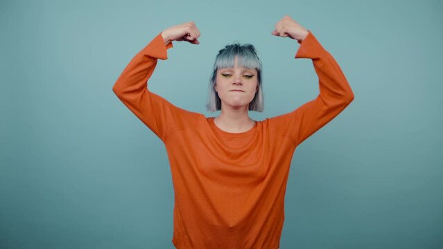 Funny hipster girl in green sweatshirt shows biceps and strength, kisses hand while standing on blue background,
