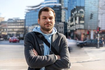 Man with arms crossed standing on street against city buildings and looking at camera.