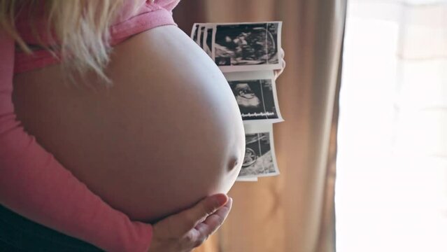 Beautiful expecting woman stroking her pregnant belly and holding ultrasound gender reveal pictures of her baby child, happy mom waiting for her beloved child, beautiful pregnant belly of attractive