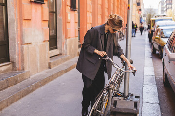 Attractive business woman come to sits on bike. Girl wear grey jacket or blazer, black pants, eyeglasses. Modern business woman ride to work on vehicle. Way to office. Riding bicycle to work.