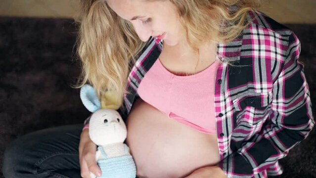 Happy smiling pregnant woman enjoying prenatal period of pregnancy at home, female sitting on the floor, holding toy and ultrasound scan pictures in hands and stroking her pregnant belly with lots of