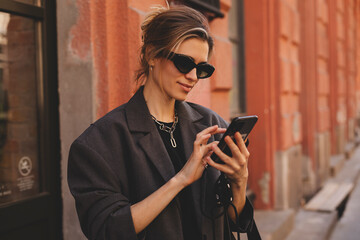 Young stylish business woman writes message on smartphone walking on old city street, hipster...
