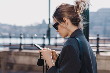 Photo of young woman dreaming, reading messages on a smartphone while going on embankment. Blonde caucasian female wear suit and sunglasses is surfing the web on a mobile phone while walking the city.