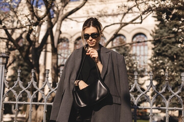 Fototapeta na wymiar Elegant young woman looking in her black leather bag her phone or purse. Business style woman wear grey blazer, black eyeglasses and bag on the street. Street style, fashion outfit.