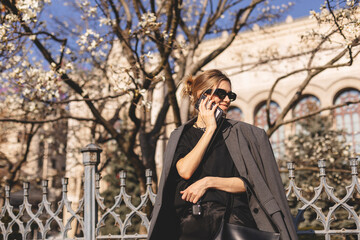 Beautiful young woman wearing suit, walking outdoors, talking on mobile phone. Beautiful Woman talking on phone. Portrait of stylish smiling business woman calling on mobile phone under blooming tree.