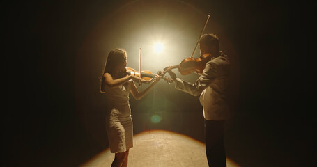 Male and female violinists performing on stage together, playing their instruments, spotted by light on black background  © andreybiling