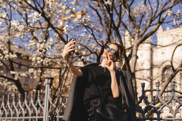 Fototapeta na wymiar Beautiful smiling blonde model in summer or spring formal clothes. Carefree female posing in sunglasses. Taking selfie self portrait photos on smartphone, shows peace sign, kiss. Focus on peace sign.