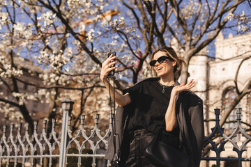 Fototapeta na wymiar Happy young woman photographing herself using her mobile phone. Caucasian female talking selfie with her smart phone on blooming tree background. Girl posing in sunglasses, suit, t-shirt, pants, bag.
