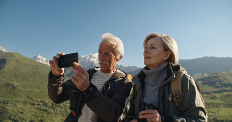 Senior caucasian couple having a nordic walk in mountains, then stopping to take a picture with smartphone. Old people travelling together after retirement - pension, tourism concept