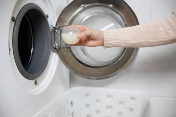Female hands pouring liquid laundry detergent into measure cup. Washing machine and clothes in white modern room