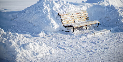 Wooden bench stands among the snowdrifts of a winter city street. Consequences of snowfall and snowstorm. Early morning.