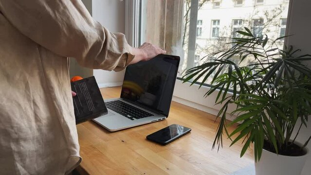 The woman closes her laptop and opens the book. She reads a book near the window. The concept of home leisure.