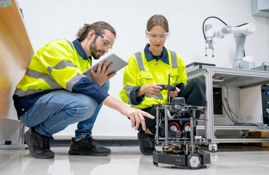 Two Caucasian Professional Technician Or Engineer Workers Sit On The Floor And Help To Check And Maintenance Small Robotic Machine In Factory Room Workplace.