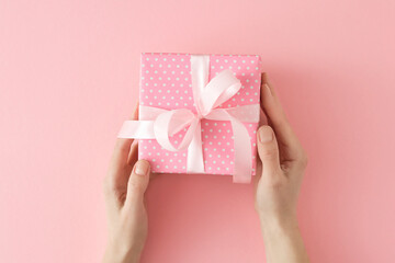 Woman day gift concept. First person top view photo of female hands holding present gift box with silk ribbon bow on pastel pink background.