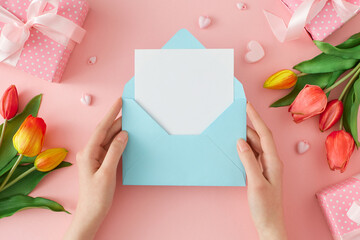 Hello spring concept. First person top view photo of female hands holding envelope with letter and tulips flowers gift boxes on pastel pink background. Holiday card idea.