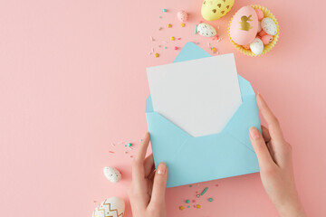 Easter concept. First person top view photo of female hands holding open envelope and yellow white gold eggs in paper basket on pastel pink background with empty space.